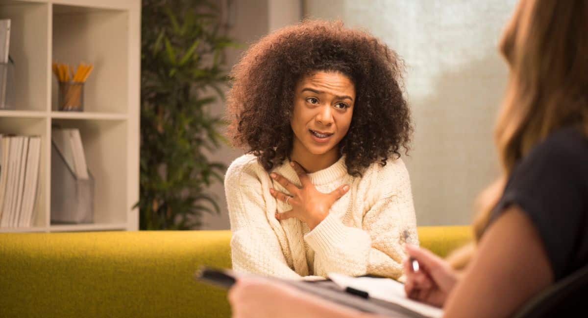 a curly woman gripping her chest with her left hand while chatting to someone